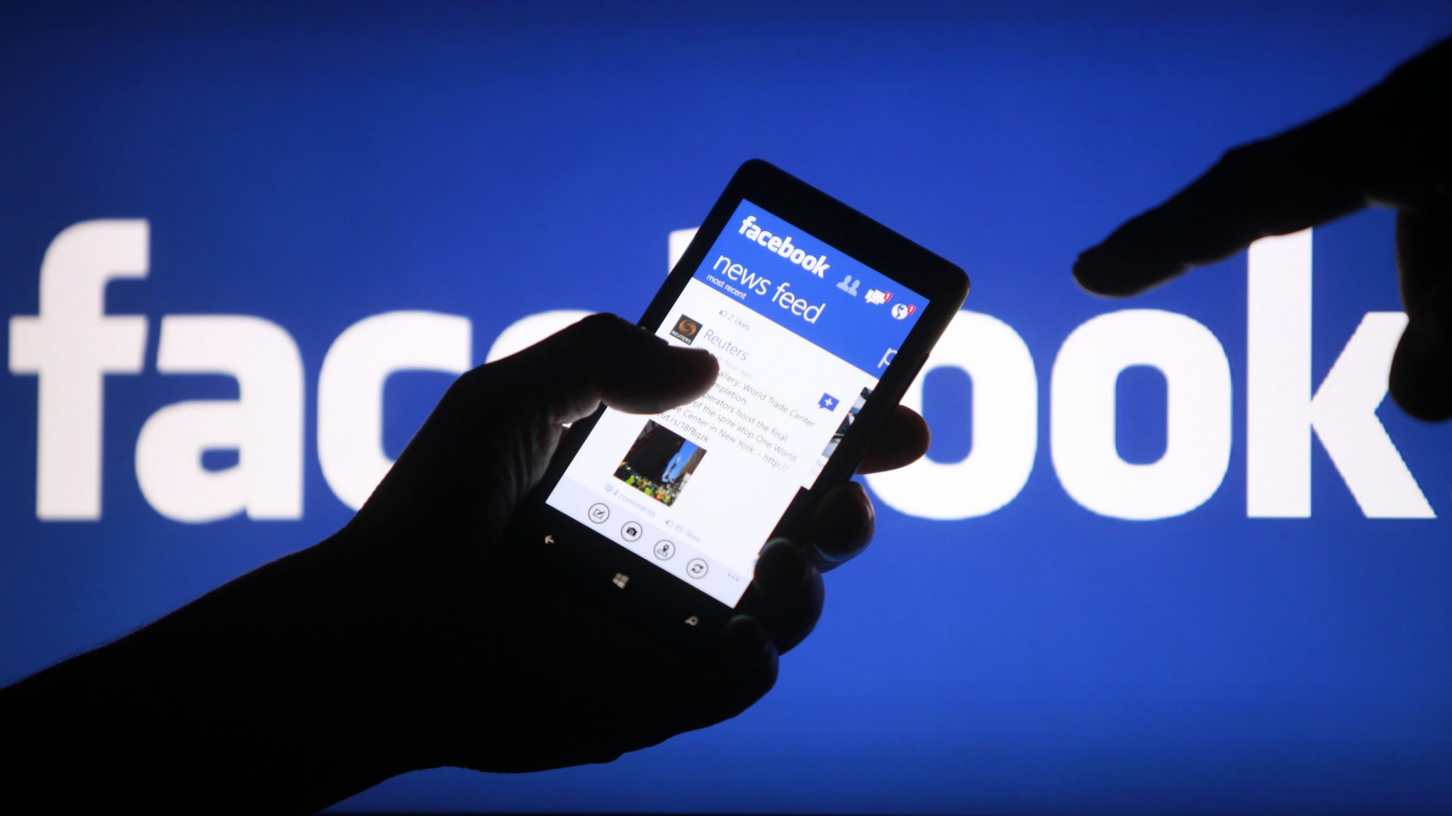 A person's hand holding a device displaying a Facebook news feed.